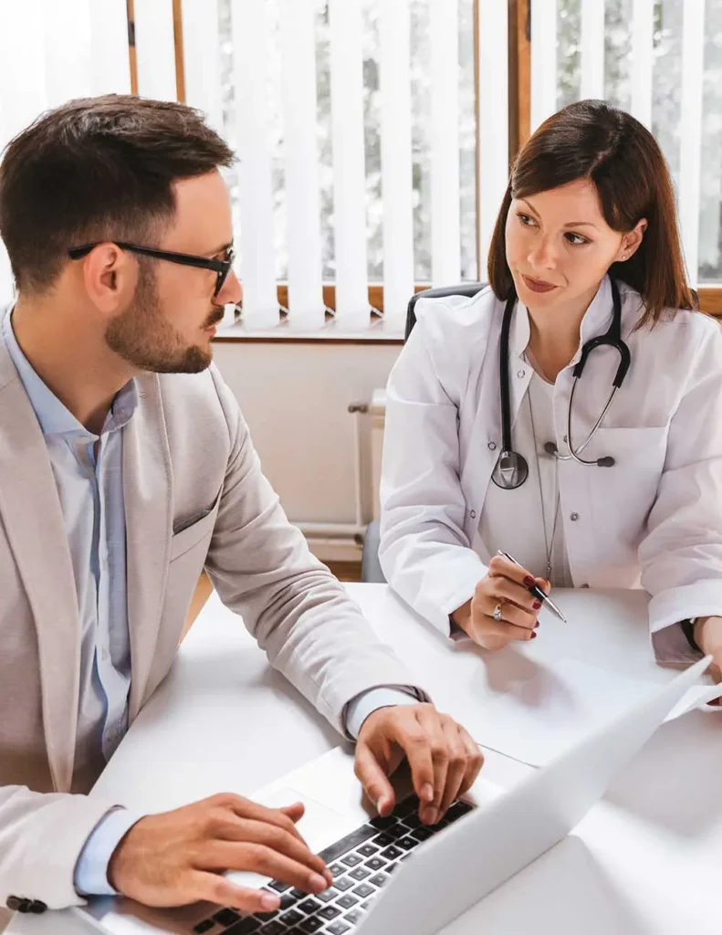 A doctor in a white coat discusses important documents about medical billing services for small practices with a professional man using a laptop in a bright office setting. The interaction between the two highlights collaboration and careful review, emphasizing attention to detail and accuracy in medical and business processes.